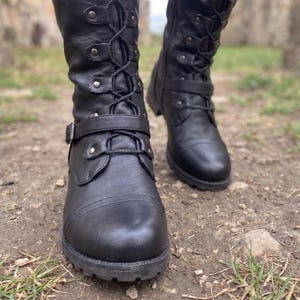 May include: A pair of black mid-calf boots featuring a lace-up front with silver-toned metal eyelets and hooks. The boots include a decorative strap across the mid-foot with a silver buckle and a rounded toe cap, finished with a thick lugged sole.