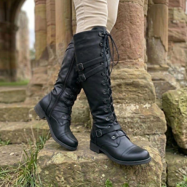 May include: Black knee-high boots featuring a lace-up front design with metal eyelets, side zippers, and two horizontal buckled straps across the shaft. The boots have a rounded toe, a low block heel, and a smooth finish on the exterior surface.