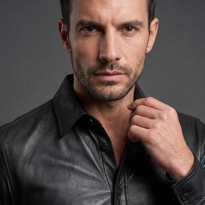 May include: A close-up portrait of a person wearing a black leather shirt with a collared neckline. The shirt is buttoned up, and the person's hand is near the collar. The background is a solid gray.