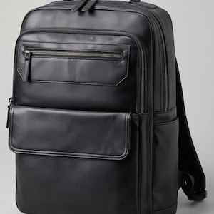 Könnte beinhalten: Ein schwarzer Rucksack mit glatter Oberfläche, einem Tragegriff oben und verstellbaren Schultergurten. Die Vorderseite verfügt über eine horizontale Reißverschlusstasche über einer größeren Klappentasche. Das Hauptfach hat einen Doppelreißverschluss.