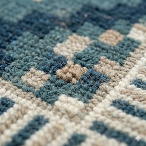 May include: Close-up of a textured rug with a geometric pattern. The rug features a combination of blue, teal, and beige tones. The design is composed of squares and diagonal lines, creating a visually interesting surface.