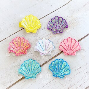 May include: Set of 8 sparkly seashell hair clips in various colors, including yellow, purple, pink, white, aqua, and blue. The clips are arranged on a white wooden surface. Olivia's Closet is written on the surface.