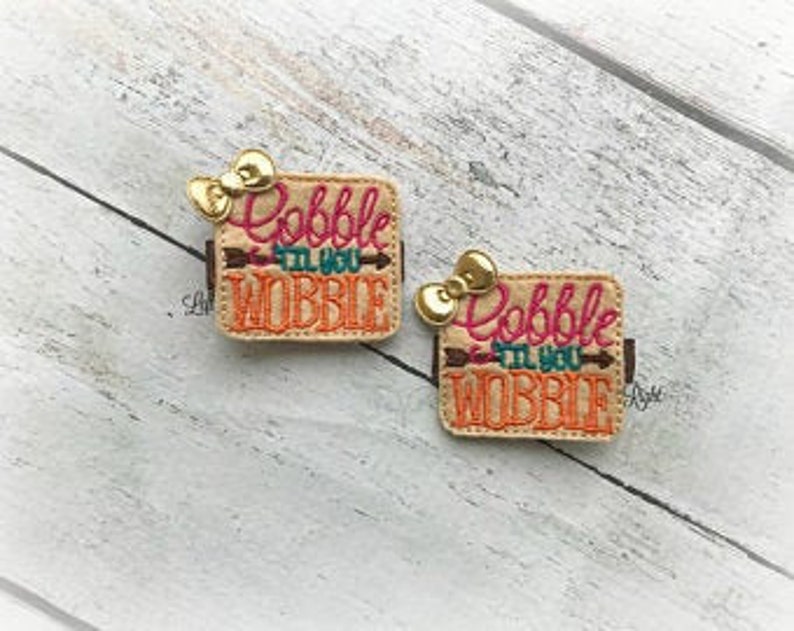 May include: Two tan felt squares with the text "Gobble 'til you Wobble" in pink, blue, and green lettering with a gold arrow. Each square has a gold bow on top.