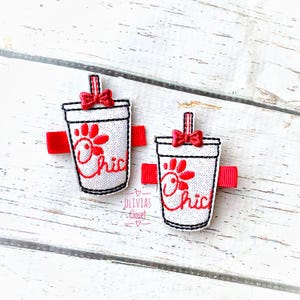 May include: Two white and red hair clips shaped like a fast food drink cup. Each clip has a red bow, a red straw, and the word "Chic" in red script. The clips are on a white wooden surface.