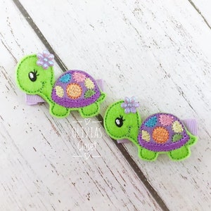 May include: Two green felt turtle hair clips with purple shells, colorful polka dots, and a single purple flower on each.