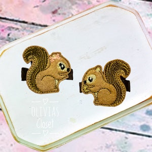 May include: Two brown embroidered squirrel hair clips with black accents. The clips are on a white background with the text "Olivia's Closet" in black.