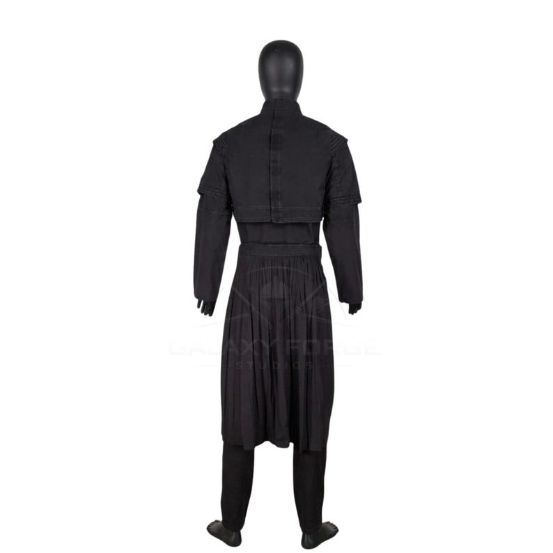 Op de afbeelding: Een zwarte set bestaande uit een tuniek met lange mouwen en een broek op een etalagepop. De tuniek heeft een hoge kraag, een gestructureerde schouderbedekking en een geplooid rokgedeelte dat tot op de knie&euml;n valt. Het kledingstuk is zwart met zichtbare stiksels.