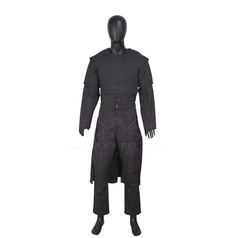 Op de afbeelding: Een zwart kostuumset op een etalagepop, bestaande uit een gestructureerd borststuk met verticale stiksels, lange mouwen en een bijpassende broek met verstevigde kniestukken en een lange, aan de voorzijde open overjas.