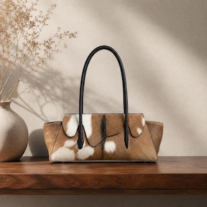 Könnte beinhalten: Eine horizontale Handtasche aus braun-weißem Rindsleder mit texturierter Felloberfläche. Die Tasche hat eine strukturierte Silhouette mit einer geschwungenen oberen Klappe und zwei hohen, dünnen schwarzen Ledergriffen an den Vorder- und Rückseiten.