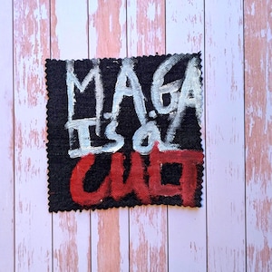 May include: A black square patch with the words "MAGA It's a Cult" in white and red paint. The patch is on a light pink and white wooden surface.