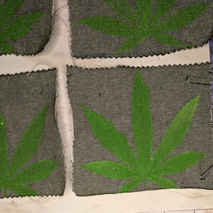 May include: Close-up of several gray fabric swatches, each featuring a vibrant green cannabis leaf design. The leaves are prominently displayed against the textured background. The edges of the fabric are unfinished.