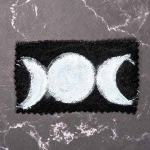 May include: A black patch featuring a white triple moon design. The design includes a full moon in the center, flanked by crescent moons on either side. The patch has a textured appearance and a decorative edge.