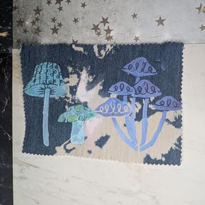 Pode incluir: Uma amostra de tecido com um desenho de cogumelos em azul, verde e roxo. Os cogumelos têm padrões detalhados. O fundo tem uma textura semelhante ao denim com um padrão claro.