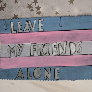 以下が含まれることがあります： 「LEAVE MY FRIENDS ALONE」の文字が銀色の手作り木製サイン。青、ピンク、グレーの横縞模様で塗装され、上部にペーパークリップが付いています。サインは装飾的なジグザグの縁取りが施されています。