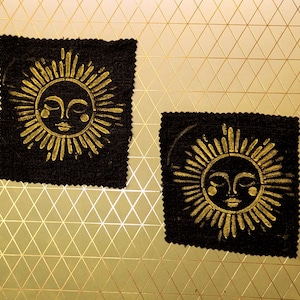 May include: Two black fabric patches with a gold sun design. The sun has a face with closed eyes and radiating lines. The patches are on a gold geometric patterned background.