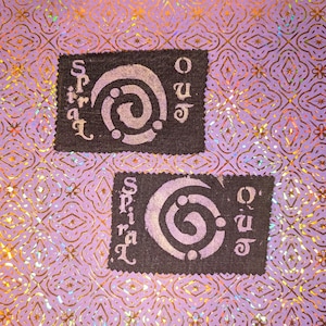 May include: Two black rectangular patches with the words "spiral" and "out" in a stylized font. Each patch features a white spiral design. The background is a purple, patterned, iridescent surface.