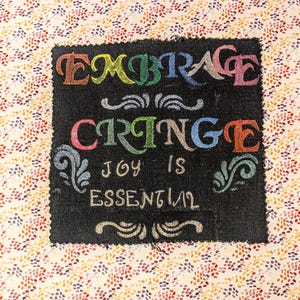 May include: A black fabric square with the words "EMBRACE CRINGE JOY IS ESSENTIAL" in colorful, stylized lettering. The background is a cream-colored fabric with a pattern of small, colorful dots.