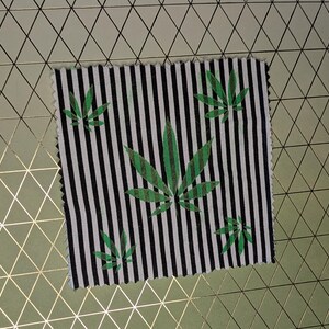 May include: A square fabric swatch with a black and white striped pattern. Green cannabis leaves are printed on the fabric. The fabric is placed on a gold and white geometric patterned surface.