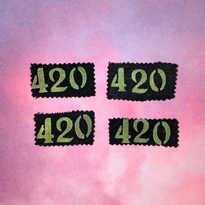 May include: Four black rectangular patches with the number "420" in green stencil font. The patches are arranged in a square on a pink and purple watercolor background. The edges of the patches have a decorative, scalloped border.