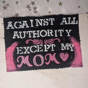 Könnte beinhalten: Schwarze rechteckige Fußmatte mit weißem Schablonentext: „AGAINST ALL AUTHORITY EXCEPT MY MOM“. Rosa Akzente und eine Herzgrafik sind ebenfalls vorhanden. Zwei Büroklammern halten die Matte an Ort und Stelle.