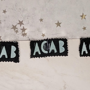 May include: Three black fabric patches with the letters "ACAB" in light blue, attached to a white surface with paper clips. The background features a textured gray surface with star-shaped cutouts.