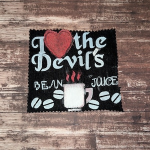 Op de afbeelding: Zwart stoffen vierkant met de tekst "I love the Devil's Bean Juice" in witte letters. Een rood hart staat boven "Devil's". Een witte koffiemok met stoom en koffiebonen zijn ook afgebeeld.