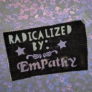 Puede incluir: Un parche rectangular negro con las palabras "RADICALIZED BY: Empathy" en letras blancas y moradas. El parche incluye dos formas de estrella y remolinos decorativos. El fondo presenta un estampado floral en tonos morados y azules.