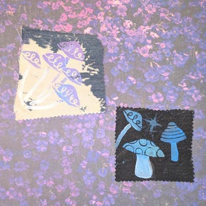 May include: Two fabric patches with mushroom designs. One patch features purple mushrooms on a cream background, the other has blue mushrooms on a black background. The background is a floral pattern in shades of purple and lavender.