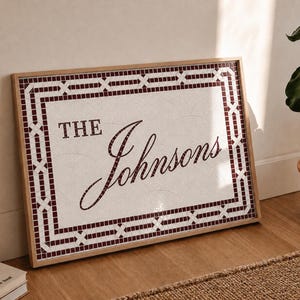 May include: A rectangular framed mosaic sign featuring a white background with a dark red geometric border. The text THE Johnsons is displayed in dark red mosaic tiles in the center of the sign, set within a light-colored wooden frame.