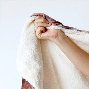May include: A close-up of a soft, plush blanket with a white, fluffy interior and a patterned exterior in shades of brown and beige. The blanket is being held up, showcasing its texture and warmth.