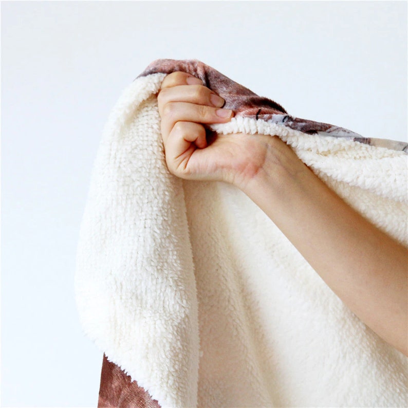 May include: A close-up of a soft, plush blanket with a white, fluffy interior and a patterned exterior in shades of brown and beige. The blanket is being held up, showcasing its texture and warmth.