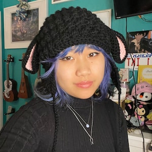 May include: A person wearing a black knit hat with pink bunny ears and a black turtleneck sweater. The hat has long strings that hang down the person's back. The person has blue hair and is wearing a silver necklace.