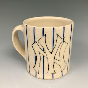 May include: Cream-colored ceramic mug with blue vertical stripes and a black and white "NY" logo. The mug has a handle and a speckled texture, suggesting a handmade quality. A sports-themed item.