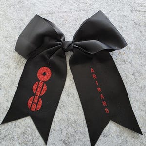 May include: Black cheerleading bow with red glitter accents. The bow features the word "ARIRANG" in red lettering on one tail and a red glitter design on the other. The bow is made of a ribbed fabric.