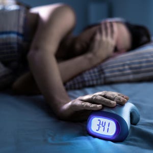 May include: A digital alarm clock displaying 3:41 sits on a bed next to a person. The person is lying in bed, with their face covered by their hand. The clock has a blue and white display.