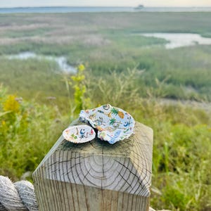 May include: Two decorative seashells featuring a white interior with a colourful pattern of small illustrations including pineapples, palm trees, sailboats, and crescent moons. The shells are resting on a wooden post with a visible grain texture.