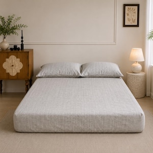 Op de afbeelding: Een bed met een lichtgrijs, gestructureerd hoeslaken in linnenlook en twee bijpassende rechthoekige kussens. Het beddengoed heeft een subtiele, geweven uitstraling met een neutrale tint, wat zorgt voor een strak en uniform oppervlak.