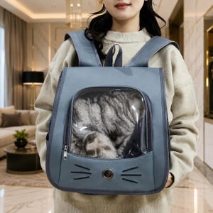 May include: A slate blue pet carrier backpack featuring a large transparent viewing window and a cat-shaped design with embroidered whiskers and a central circular ventilation grommet. The backpack has adjustable shoulder straps and a zipper closure.
