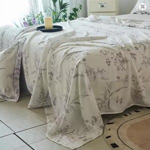 May include: A white bed sheet featuring a delicate, repeating botanical print in a soft grey tone. The fabric drapes over the edge of a bed, showcasing intricate illustrations of various wildflowers, grasses, and leafy stems across the surface.