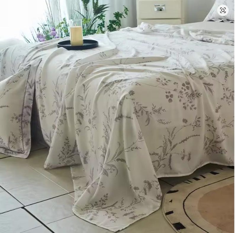 May include: A white bed sheet featuring a delicate, repeating botanical print in a soft grey tone. The fabric drapes over the edge of a bed, showcasing intricate illustrations of various wildflowers, grasses, and leafy stems across the surface.