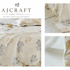 May include: A cream-coloured bed sheet set featuring a repeating botanical pattern of small blue and gold leaves. The text AJCRAFT HOME TEXTILE BUTTERFLY DANCE AND CLEAR BREEZE is printed at the top. The fabric displays a soft texture with visible stitching along the edges of the pillowcases and the main sheet.