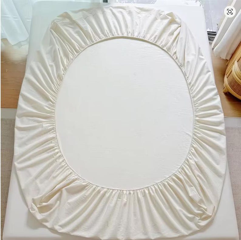 May include: An oval-shaped fitted sheet in a solid off-white color. The sheet features an elasticized border with gathered fabric detailing around the entire perimeter, designed to fit snugly over an oval mattress surface.