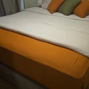 Op de afbeelding: Een bed met een effen mosterdgele bedrok en een bijpassend mosterdgeel laken over het matras. Het beddengoed bestaat uit een wit dekbedovertrek en een verzameling decoratieve kussens in mosterdgeel en olijfgroen.