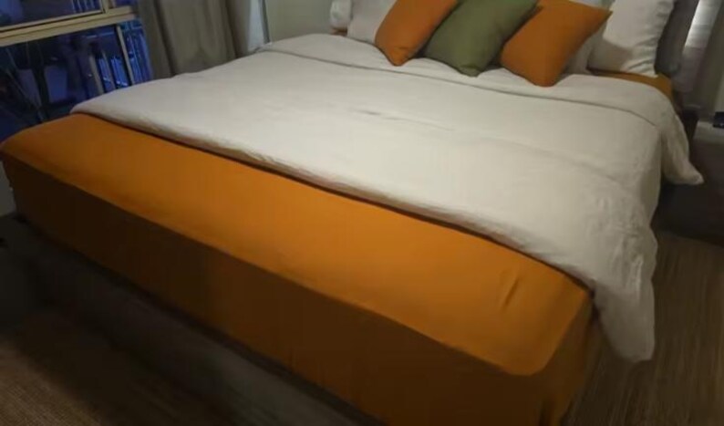 Op de afbeelding: Een bed met een effen mosterdgele bedrok en een bijpassend mosterdgeel laken over het matras. Het beddengoed bestaat uit een wit dekbedovertrek en een verzameling decoratieve kussens in mosterdgeel en olijfgroen.