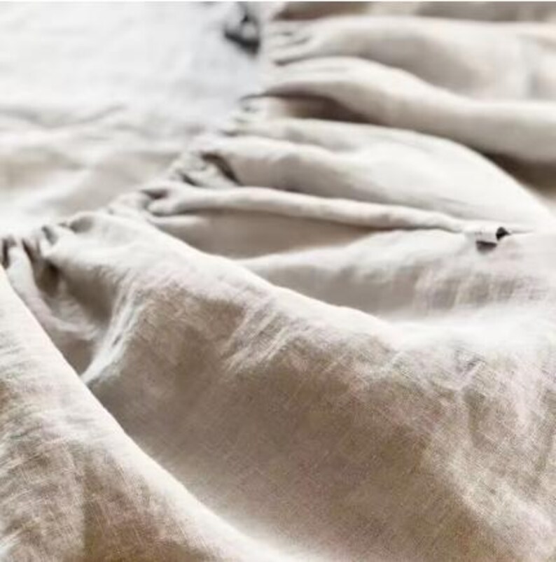 Op de afbeelding: Een lichtbeige linnen laken met een geplooide, gerimpelde rand en een zichtbare elastische naad. De stof heeft een natuurlijke, getextureerde weving en een zachte, matte afwerking, waarbij de plooien en de valling van het materiaal opvallen.