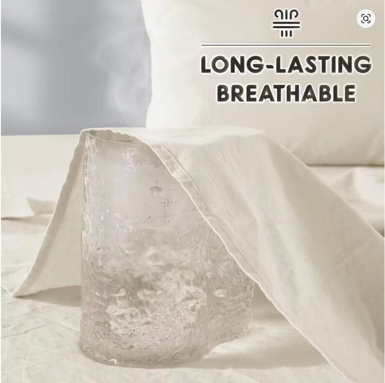 May include: A clear, textured glass tumbler is partially covered by a piece of off-white fabric. Above the fabric, the text "LONG-LASTING BREATHABLE" is displayed in dark brown letters, accompanied by a small icon of three horizontal lines.