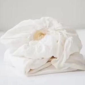 Op de afbeelding: Een opgevouwen, off-white hoeslaken gemaakt van een zachte, geweven stof. Het laken heeft een elastische rand die aan de bovenkant is geplooid, wat zorgt voor een ronde, gebundelde uitstraling met zichtbare vouwen en zachte schaduwen.