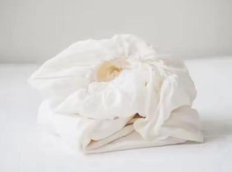 Op de afbeelding: Een opgevouwen, off-white hoeslaken gemaakt van een zachte, geweven stof. Het laken heeft een elastische rand die aan de bovenkant is geplooid, wat zorgt voor een ronde, gebundelde uitstraling met zichtbare vouwen en zachte schaduwen.
