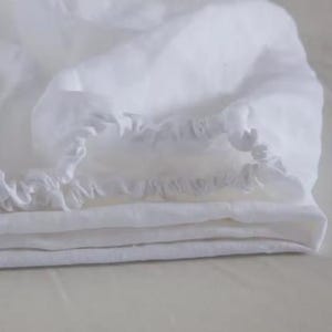 Op de afbeelding: Een wit hoeslaken met een geplooide elastische rand langs de omtrek. De stof heeft een zacht, licht getextureerd uiterlijk met zichtbare vouwen en lagen, wat de heldere, witte kleur van het textielmateriaal goed laat uitkomen.