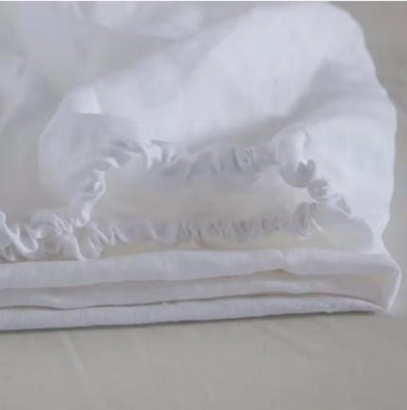 Op de afbeelding: Een wit hoeslaken met een geplooide elastische rand langs de omtrek. De stof heeft een zacht, licht getextureerd uiterlijk met zichtbare vouwen en lagen, wat de heldere, witte kleur van het textielmateriaal goed laat uitkomen.